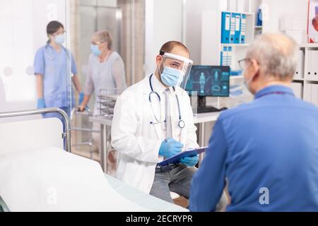 Doctor wearing visor as safety precaution in the course of consultation of senior man. Physician practitioner in the course of private modern clinic appointment during coronavirus pandemic. Stock Photo