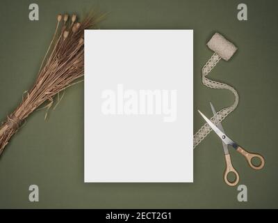 Invitation card mockup on green background. Blank white card with dried wild poppies, scissors and textured beige lace. Workspace concept. Overhead. Stock Photo