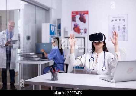 Caucasian female physician in white gown and VR headset sitting at desk in cabinet swiping and scrolling with hands in air. Virtual reality concept. Healthcare futuristic. Doctor working in future. Stock Photo