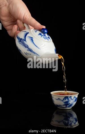 Woman's hand serving tea with blue china teapot in cup, on black background, with reflection, vertical, with copy space Stock Photo