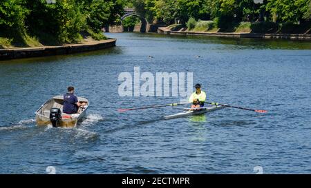 2 men in boats on sunny scenic River Ouse (sculler rowing single scull boat or shell & man in motorboat coaching) - York, North Yorkshire, England UK. Stock Photo