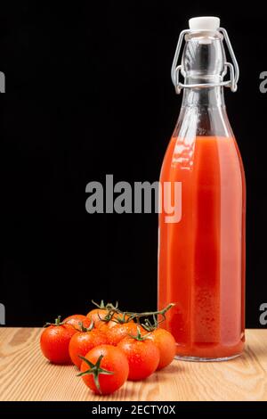 View of glass bottle with tomato juice on wooden table with cherry tomatoes, black background, vertical, with copy space Stock Photo