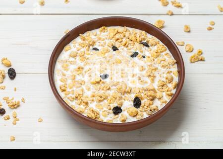 Healthy breakfast. Muesli with dried grapes is poured with milk in a clay bowl on a white wooden table. Muesli is scattered on the table. Space for te Stock Photo