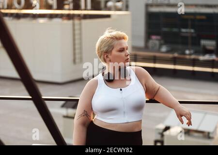 Woman with oversized body leaning to a railing during a workout on a roof. Oversize fitness woman in sportswear wearing headphones looking away outdoo Stock Photo