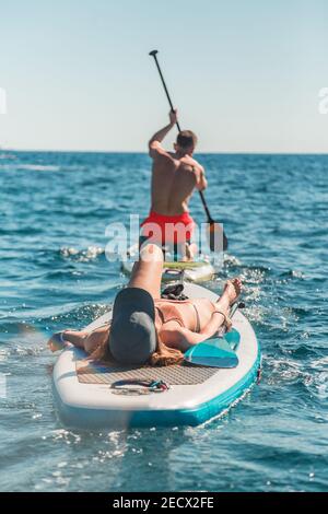 A vertical shot of a beautiful woman sleeping on SUP paddleboard and a man rowing in the Adriatic sea Stock Photo