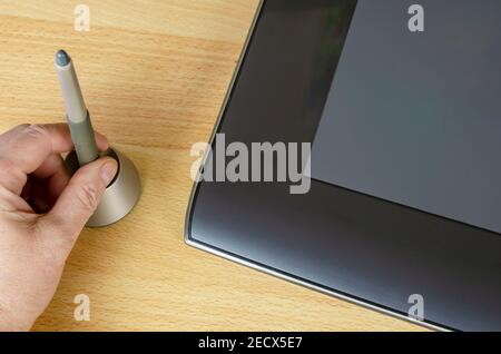 A man's hand retrieves a pen for a graphic tablet from a special stand. A professional device for designers lies next to it on the table. An angled vi Stock Photo