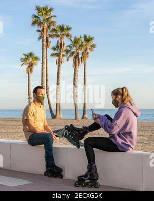 Roller skating couple in medical protective mask (against coronavirus , covid-19)sitting on promenade,using cell phone on seaside, palm trees and beac Stock Photo