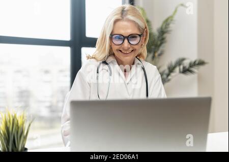 Female doctor with blond hair and stethoscoper,in black glasses sitting at the laptop chatting wit patient or family,smiling,at the world online Stock Photo