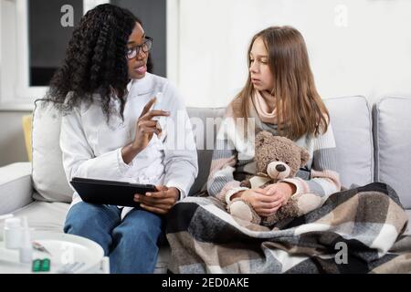 Medicines prescribing and appointment. Female African pediatrician visiting her sick teen girl patient at home, and giving directions how to use nasal Stock Photo