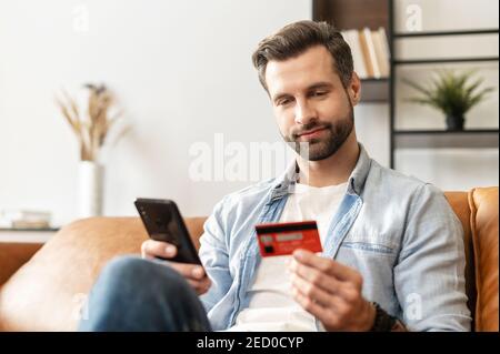 Bearded handsome man makes purchase online. A hipster wearing casual shirt holds a mobile phone and a credit card orders food. Smiling guy is using Stock Photo