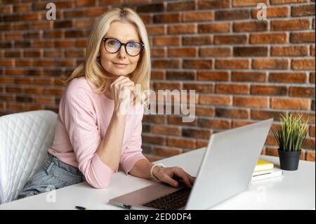 Beautiful mature businesswoman working with laptop,looking at the camera, smiling and typing, interrupted from work,best worker,workoholic,beautiful Stock Photo