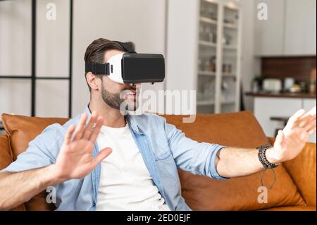 Excited young bearded man wearing glasses of virtual reality sitting on the comfortable couch at home, a guy in VR headset lost in cyberspace, using Stock Photo