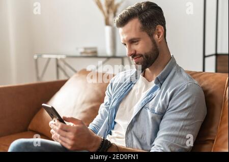 Smiling guy wearing casual outfit messaging, texting on the smartphone. Side view a young man sits on the modern sofa and using mobile app, scrolling Stock Photo