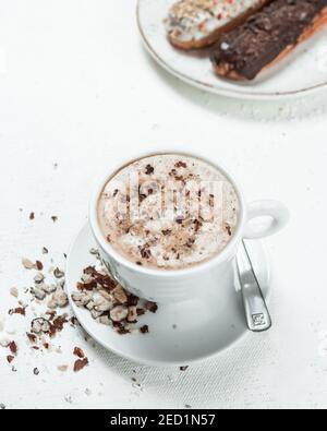 A vertical shot of Cappuccino with hazelnuts and eclair on a white table Stock Photo
