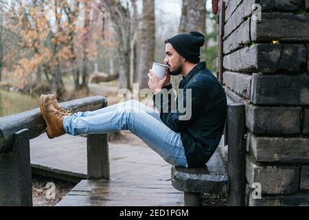 A young hipster sitting on the terrace bench with his legs leaning against a wooden pole and drinking coffee melancholic autumn mood Stock Photo