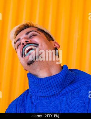 Androgynous cheerful male with blue lips and in stylish wear standing on background of yellow wall in city with eyes closed Stock Photo