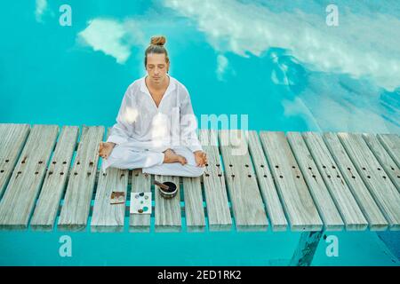 From above blond male hipster yogi in white clothes sitting in lotus pose meditating with closed eyes near tibetan singing bowl and crystals on wooden Stock Photo