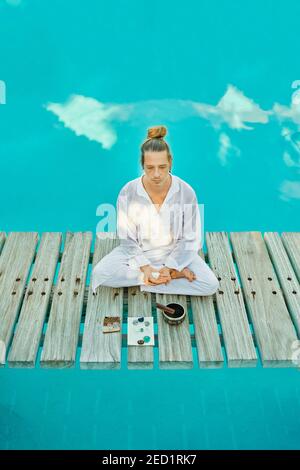 From above blond male hipster yogi in white clothes sitting in lotus pose meditating near tibetan singing bowl and crystals on wooden path bridge on t Stock Photo
