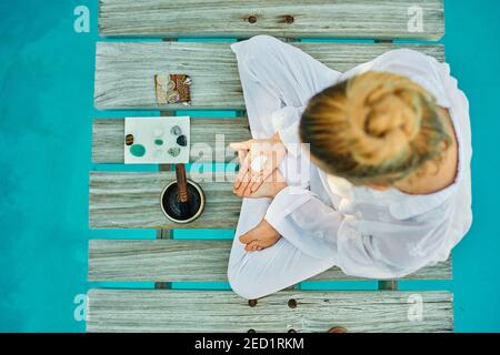 From above unrecognizable blond male hipster yogi in white clothes sitting in lotus pose meditating near tibetan singing bowl and crystals on wooden p Stock Photo