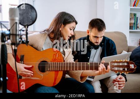 Couple of musicians sitting on sofa and recording song while playing acoustic guitar and using laptop Stock Photo
