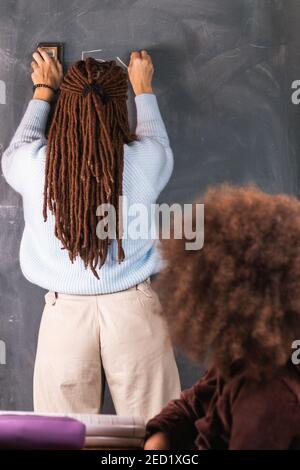 Back view of female teacher in dreadlocks writing mathematical example on chalkboard for black girl sitting at desk in classroom during lesson at scho Stock Photo