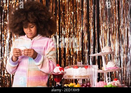 Charming African American girl with Afro hairstyle standing with donut in room with various sweets on background of golden tinsel stripes Stock Photo
