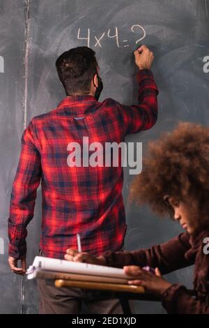 Teacher writing mathematical example on chalkboard for black girl sitting at desk in classroom during lesson at school Stock Photo