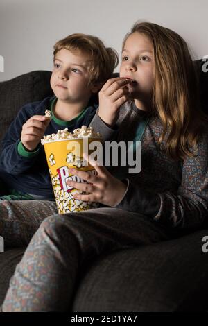 Young amazed siblings kids sitting comfortable on sofa with popcorn bucket while eating and watching TV Stock Photo