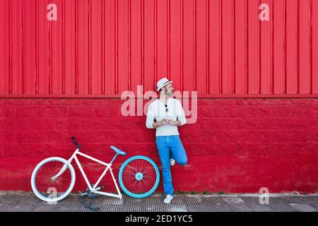 Full body of thoughtful trendy bearded male in stylish outfit leaning on red wall using mobile phone while relaxing on street near modern bicycle look Stock Photo