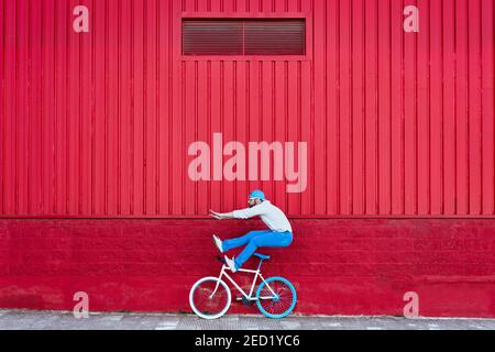 Side view of active bearded male cyclist in stylish outfit playing near bike on street near red building wall Stock Photo