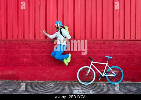 Side view of excited bearded male cyclist with backpack listening to music on headphones jumping with mouth open and fist up with victory gesture near Stock Photo
