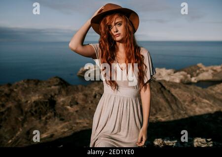 Beautiful young redhead unemotional Woman on summer dress and hat standing relaxing at mountain seaside landscape looking at camera with waving hair o Stock Photo