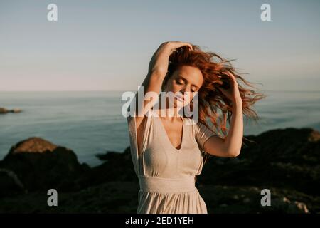 Beautiful young redhead unemotional Woman standing relaxing at mountain seaside landscape with eyes closed and waving hair on background of blue sea Stock Photo