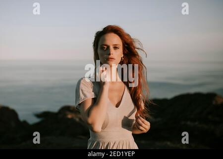 Beautiful young redhead unemotional Woman on summer dress standing relaxing at mountain seaside landscape looking at camera with waving hair on backgr Stock Photo