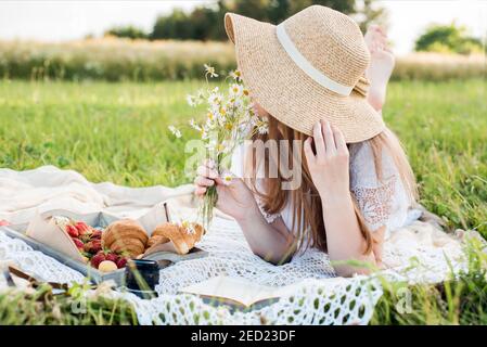 Summer day, picnic in the village. A beautiful girl in a hat lies on a plaid plaid. a bouquet of daisies, flowers in her hair. Stock Photo