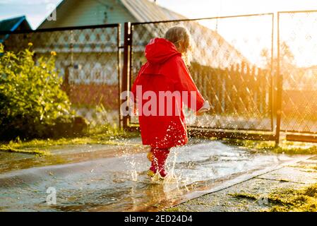 Cute girl in a red jacket is jumping in the puddle.The setting warm summer or autumn sun. summer in the village. Stock Photo