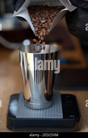 pouring coffee beans from pack into metal glass on black acaia scales in coffee shop, closeup Stock Photo
