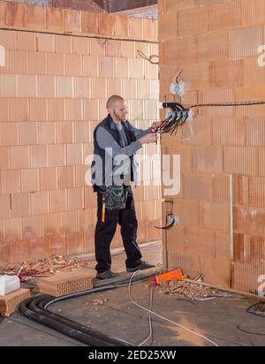 An electrician at work is prepared to install sockets in unfinished house built of clay block bricks. Worker ready for wiring cables inside house unde Stock Photo