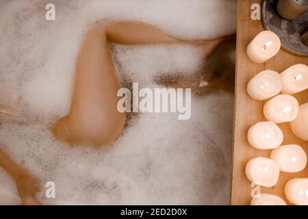 Close up photo of woman's legs in spa bathtub with foam and wooden table vase of plant candle. Modern spa center Stock Photo