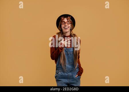 Photo of attractive female farmer looks unexpectedly surprised, wears funny glasses. White woman wears denim overall and hat isolated brown color Stock Photo