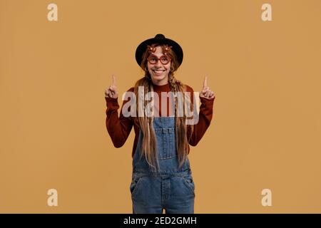 Photo of attractive female farmer looks pleasant and happy, wears funny glasses points up. White woman wears denim overall and hat isolated brown Stock Photo