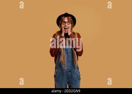 Photo of attractive female farmer looks unexpectedly surprised, wears funny glasses. White woman wears denim overall and hat isolated brown color Stock Photo