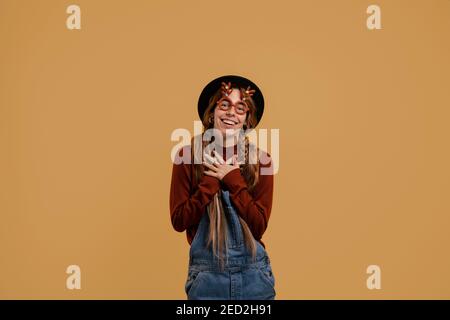 Photo of attractive female farmer looks pleasant and happy, wears funny glasses. White woman wears denim overall and hat isolated brown color Stock Photo