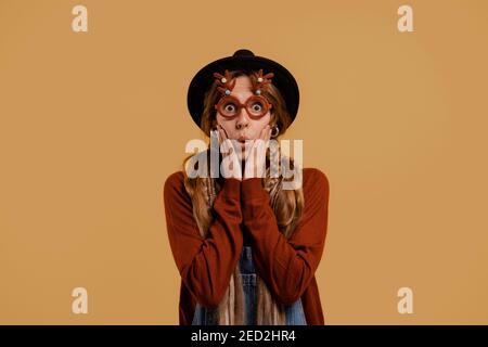Photo of attractive female farmer stands, wears funny glasses and looks very surprised. White woman wears denim overall and hat isolated brown color Stock Photo