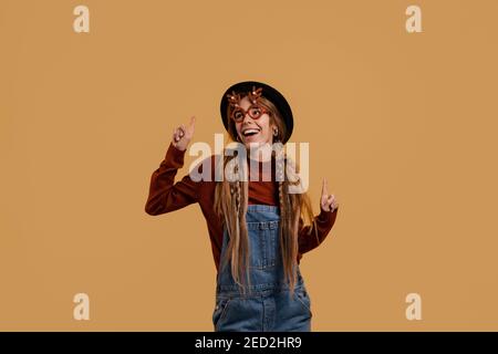 Photo of attractive female farmer dances, wears funny glasses and looks very cute. White woman wears denim overall and hat isolated brown color Stock Photo