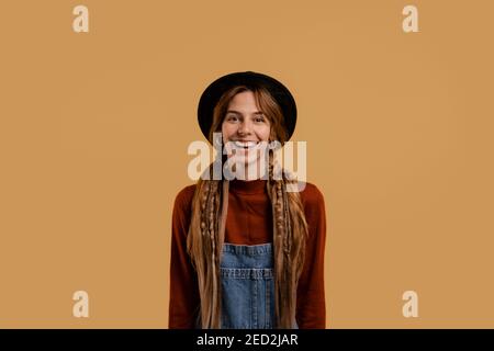Photo of attractive female farmer stands and smiles, looks so cute. White woman wears denim overall and hat isolated brown color background Stock Photo