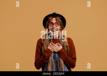 Photo of attractive female farmer stands, wears funny glasses and looks very surprised. White woman wears denim overall and hat isolated brown color Stock Photo