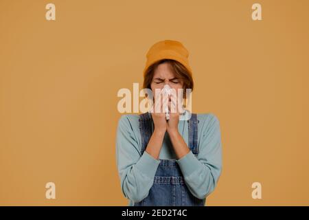 Photo of cute female farmer has got running nose and looks sick. Wears denim overalls and hat, isolated brown color background Stock Photo