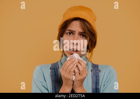 Photo of cute female farmer has got running nose and looks sick. Wears denim overalls and hat, isolated brown color background Stock Photo