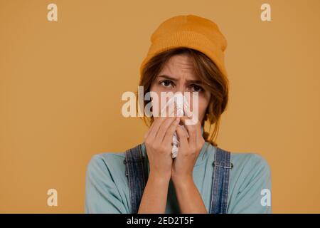 Photo of cute female farmer has got running nose and looks sick. Wears denim overalls and hat, isolated brown color background. Stock Photo